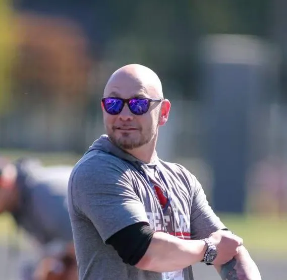 Portrait of mature man with white beard smiling outdoors on urban street with parked cars and buildings in the background.